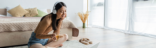 happy asian woman in headphones sitting on floor with cup of tea near laptop, banner