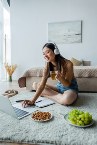 smiling asian woman with cup of tea using laptop on floor in bedroom
