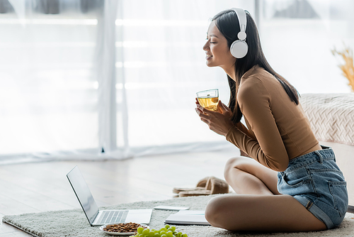happy asian woman in headphones holding cup of tea near laptop and snacks on floor