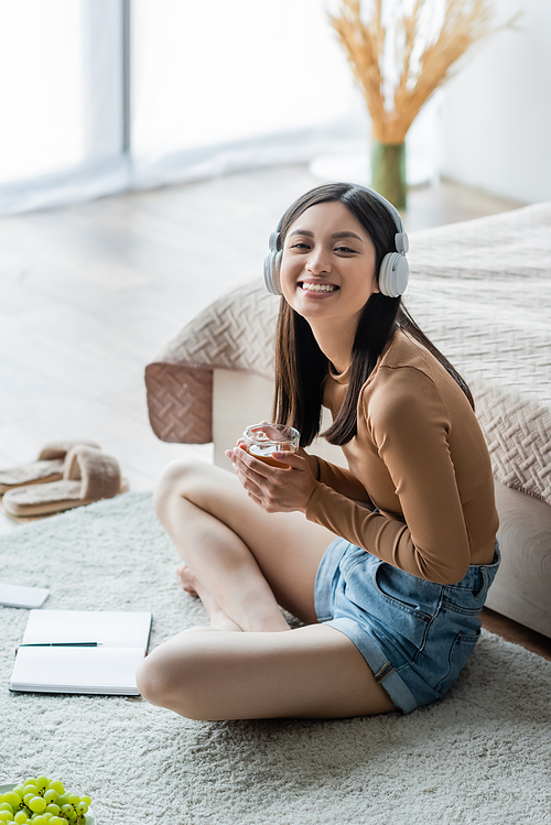 cheerful asian woman in headphones holding cup of tea while sitting on floor with crossed legs near notebook