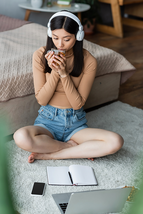 asian woman in headphones drinking tea near laptop and notebook on floor