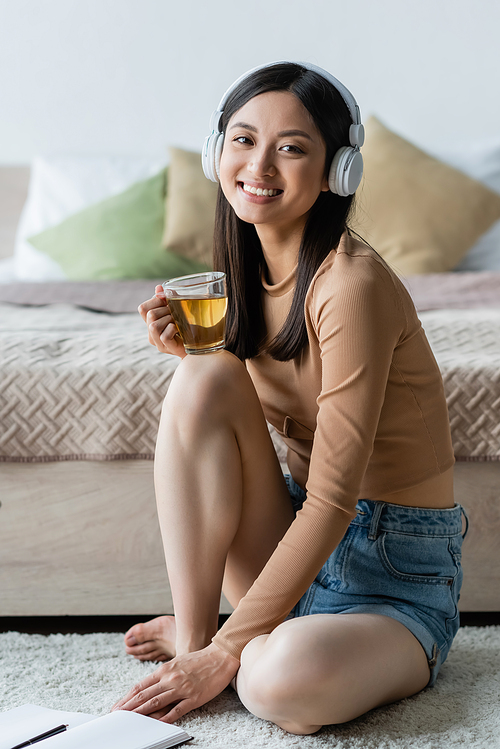 asian woman in headphones holding cup of tea and smiling at camera in bedroom