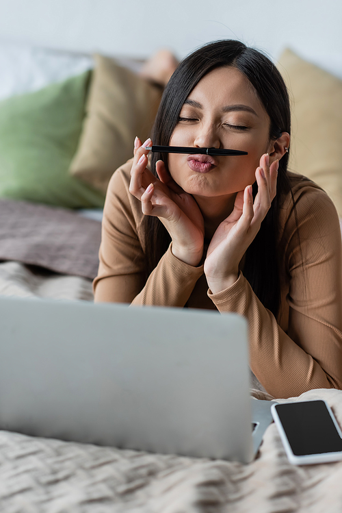 playful asian woman with pen between nose and lips lying on bed near blurred laptop and smartphone