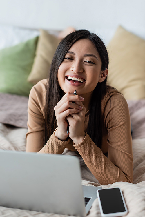 asian woman smiling at camera near blurred smartphone and laptop on bed