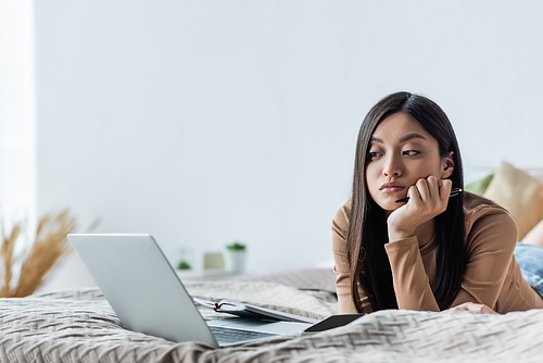 pensive asian woman looking at laptop while lying in bedroom