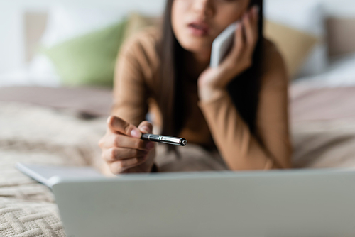 blurred woman pointing with pen at laptop while talking on cellphone in bedroom