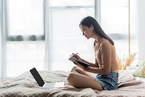 side view of asian woman sitting on bed with crossed legs and writing in notebook near laptop
