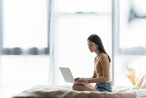pretty asian woman sitting with crossed legs on bed near large window and typing on laptop