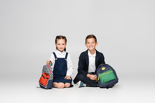 cheerful schoolkids in uniform sitting with backpacks on grey