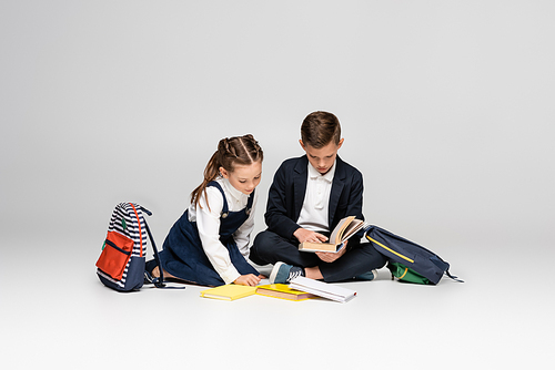schoolkids in uniform sitting and reading books near backpacks on grey