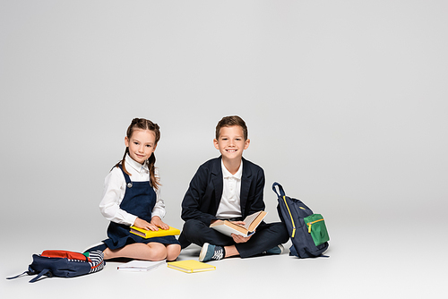 happy schoolkids in prestigious uniform sitting with backpacks and books on grey