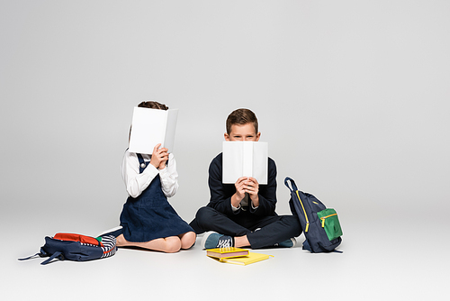 schoolkids in uniform sitting and covering faces with books near backpacks on grey