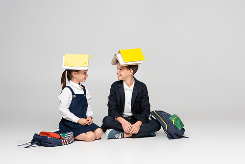 happy schoolkids in uniform with books on heads sitting and looking at each other on grey