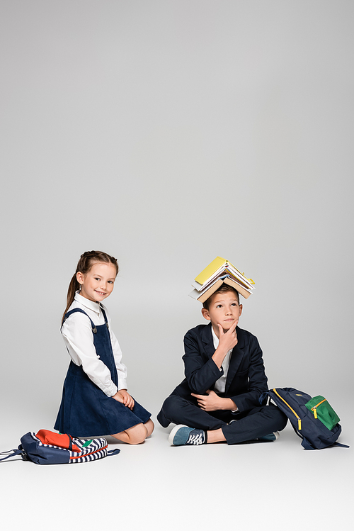 pensive schoolboy with books on head sitting near cheerful girl on grey