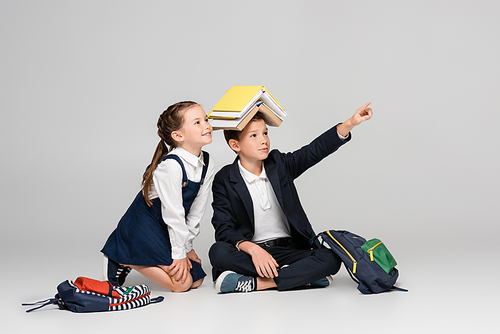 schoolboy with books on head pointing away with finger near cheerful girl on grey