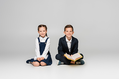 positive schoolkids sitting with books and smiling on grey