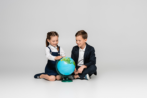 happy schoolgirl and schoolboy looking at globe while sitting on grey