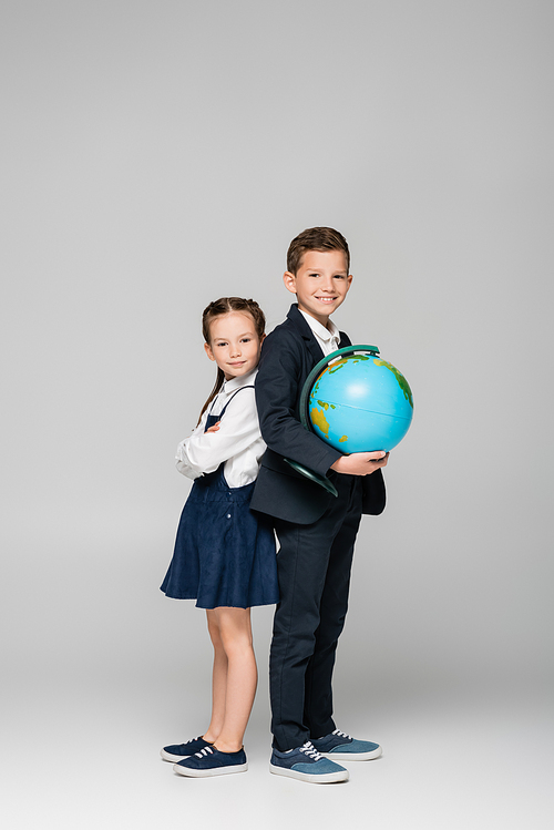happy schoolboy holding globe while standing with girl in dress on grey
