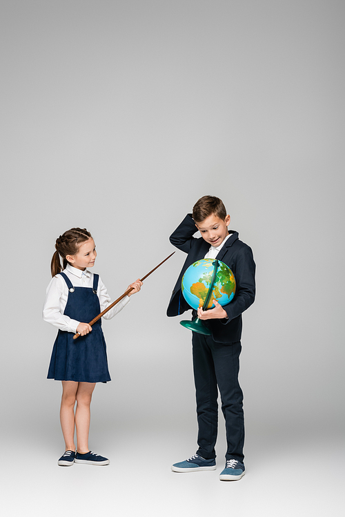 smiling schoolgirl holding pointing stick near confused boy with globe on grey
