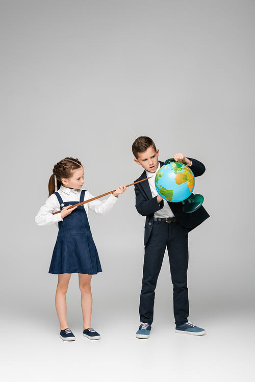 schoolgirl holding pointing stick near boy with globe on grey