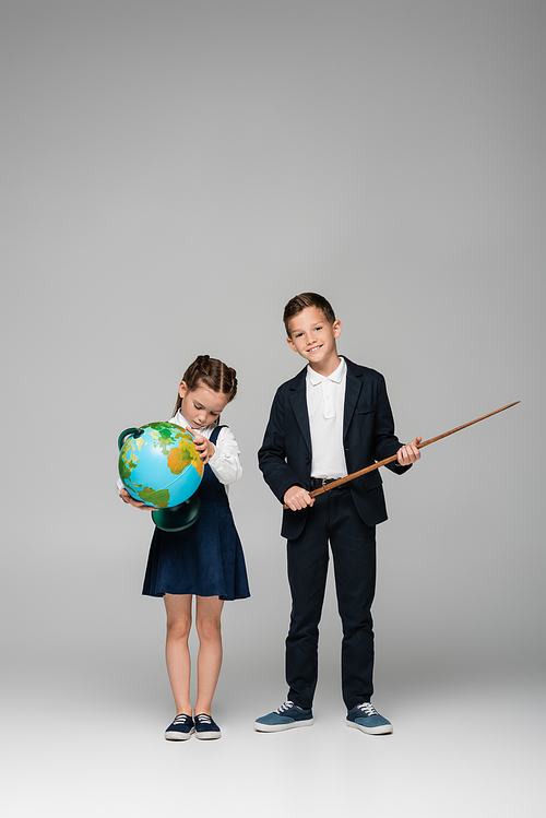 cheerful schoolboy holding pointing stick near sad girl in dress with globe on grey
