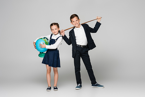 happy schoolboy holding pointing stick near smiling girl in dress with globe on grey