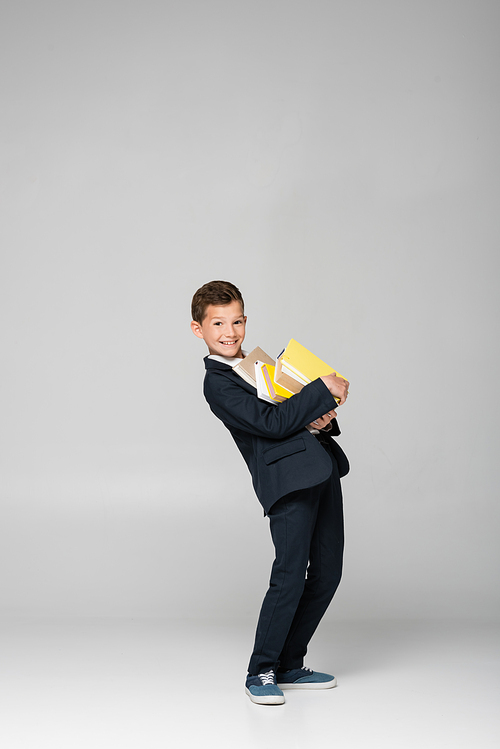 happy schoolboy in blazer and pants holding pile of books on grey