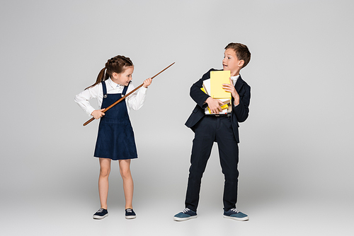 smiling schoolgirl holding pointing stick and threatening schoolboy in uniform with pile of books on grey