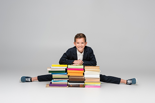 flexible schoolboy doing split near pile of books on grey