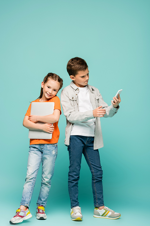 happy girl with laptop standing near boy using smartphone on blue