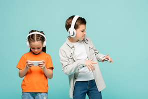 girl using smartphone near boy in wireless headphones listening music while dancing isolated on blue