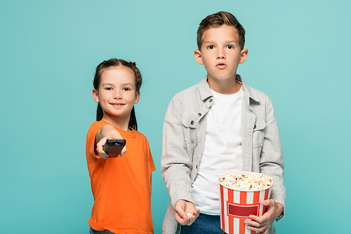 girl holding remote controller near boy with popcorn bucket isolated on blue
