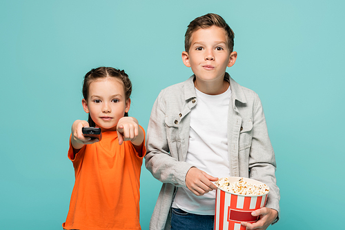 girl holding remote controller and pointing with finger near boy with popcorn bucket isolated on blue
