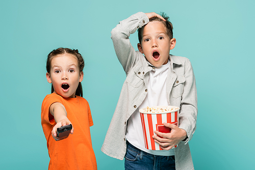 shocked girl holding remote controller near boy with popcorn bucket isolated on blue
