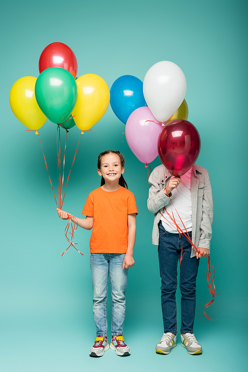 happy girl holding colorful balloons near boy obscuring face on blue