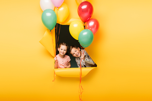 cheerful kids holding colorful balloons through hole on yellow background