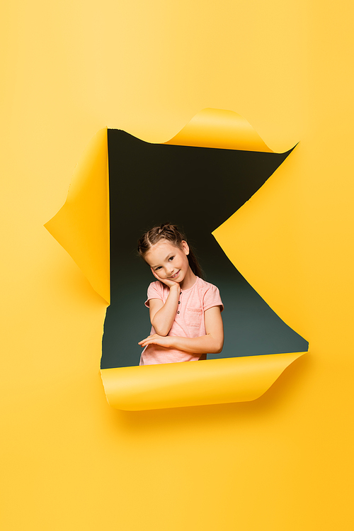 pleased kid  while smiling through ripped hole on yellow background