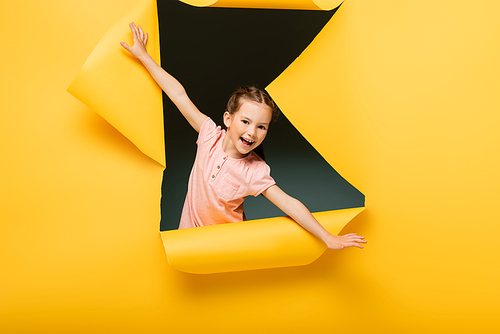 excited kid with outstretched hands  through ripped hole on yellow background