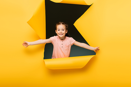 joyful kid with outstretched hands  through ripped hole on yellow background