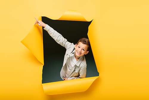 happy boy pointing with finger and  through ripped hole on yellow background