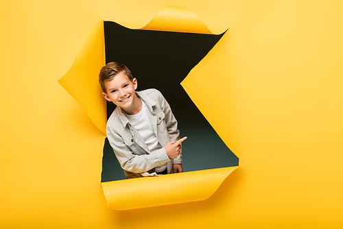 joyful boy pointing with finger and  through ripped hole on yellow background