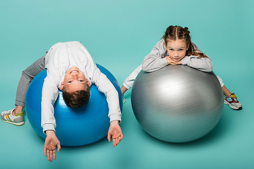 happy boy in sportswear lying on fitness ball near girl on blue