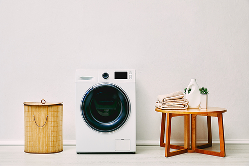 modern washing machine near laundry basket, coffee table with towels, detergent bottle and plant in bathroom
