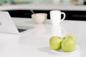 selective focus of green apples near laptop, bowl and cup on table