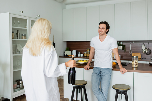 happy man looking at blonde woman with bottle of wine in kitchen