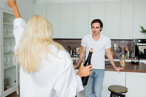 tattooed man looking at blurred blonde woman with bottle of wine in kitchen