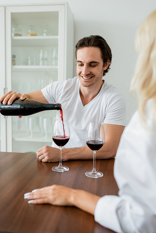 cheerful man pouring red wine into glass near blurred girlfriend