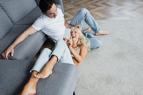 cheerful woman laughing and using smartphone while lying near boyfriend in living room