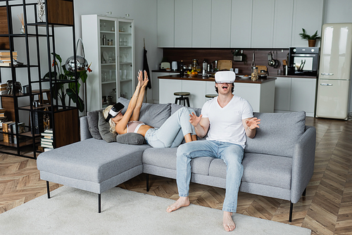 blonde woman in vr headset lying on sofa with outstretched hands near astonished boyfriend