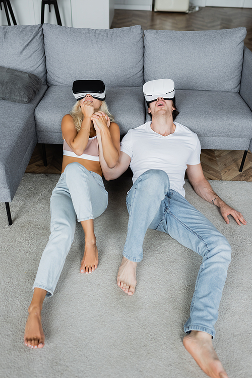shocked couple in vr headsets sitting on carpet near grey couch